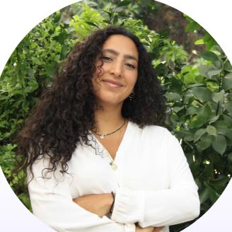 Portrait of Tala standing in front of green foliage, arms crossed, wearing a white shirt.